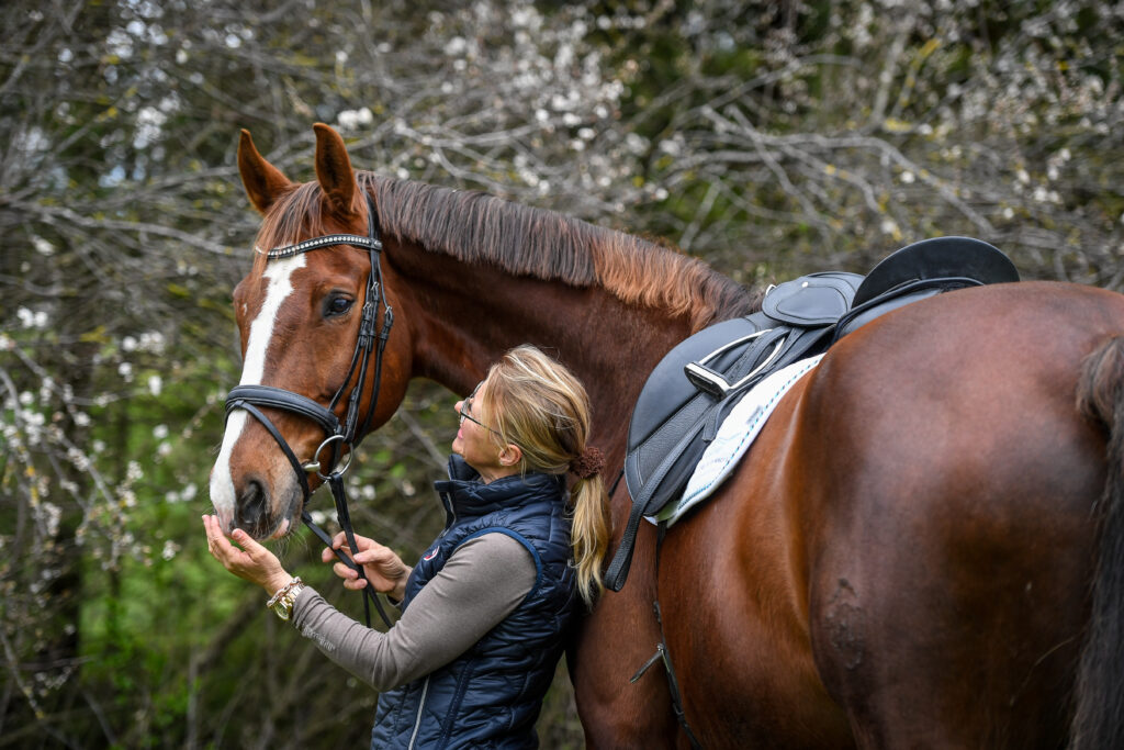 Anne Schmatelka steht neben einem gesattelten rotbraunen Pferd mit weisser Blesse, schaut zu ihm hoch und hält sein Maul in der Hand